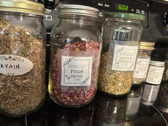 Jars of dried herbs and flowers on a kitchen countertop, including labels for 'Slirvain,' 'Rose,' and 'Yarrow,' showcasing various herbal ingredients.