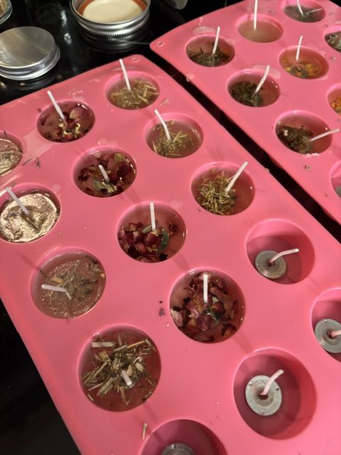 A pink silicone mold filled with various candle wax mixtures, each containing different dried herbs and flowers, with wicks protruding from the wax.