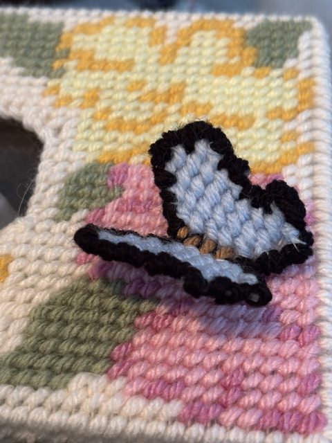 Close-up of a hand-stitched butterfly in blues and blacks on a colorful needlepoint background featuring floral patterns.