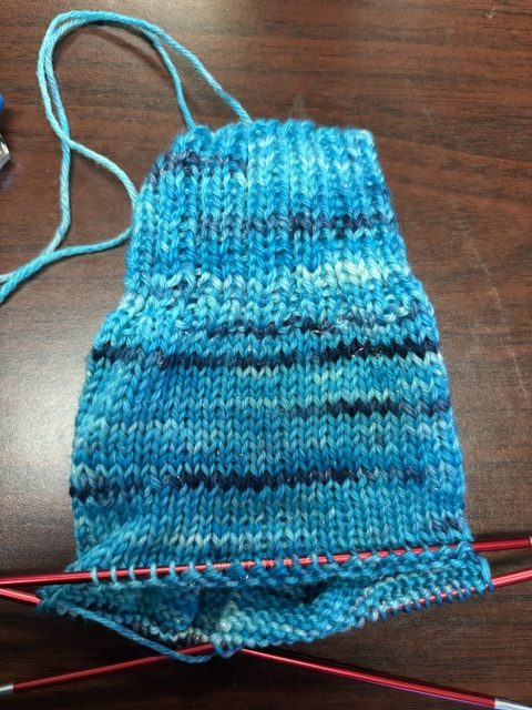A partially knitted piece of fabric in various shades of blue, showcasing a ribbed pattern with straight knitting needles resting below.