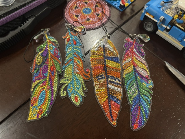 Four colorful feather-shaped keychains adorned with rhinestones, displayed on a wooden table. The feathers feature intricate designs in various vibrant colors.