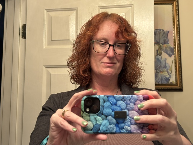 A woman with curly, red hair taking a selfie in a bathroom mirror, wearing glasses and a gray sweater. She has green nail polish and is holding a colorful phone case.