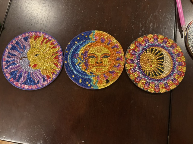 Three circular coasters featuring colorful bead designs; the first depicts a sun with a face, the second shows a stylized sun and stars, and the third features a sun motif with a dark background.