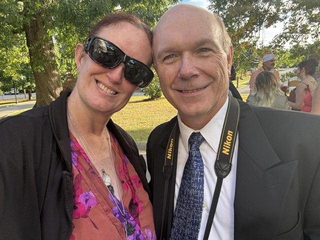 A woman smiling with sunglasses and a man in a suit with a camera around his neck, posing together outdoors.
