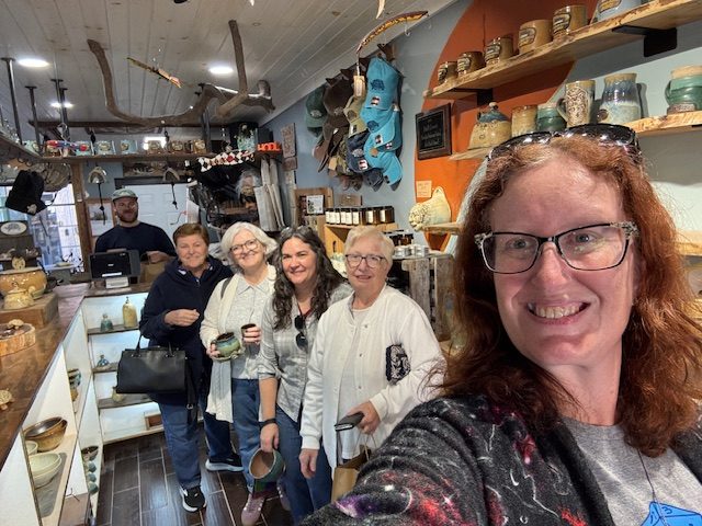 A group of six people, including a woman taking a selfie, inside a cozy shop filled with pottery and handmade items.